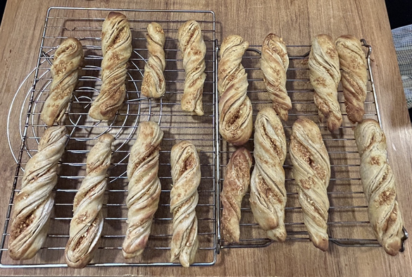 Closeup from above of 16 large cheese twisters, cooling off on my two biggest cooling racks. They are nicely golden, and the cheese is half-seeping through the places where the dough was twisted. I dusted the cheese with tandoori powder, before folding the dough and enclosing cheese & tandoori powder, after which I cut the flattish slab of dough into 16 pieces and twisted each of them.