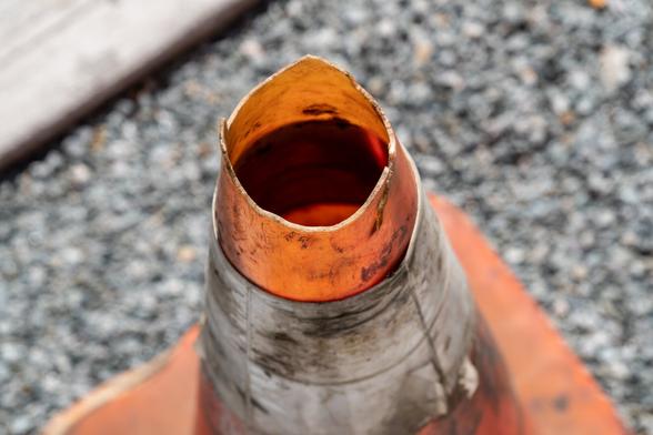 A photograph of a batter r traffic cone taken from above. The white stripe around the cone is dirty, the top is open with an uneven edge. Pavement is visible, as is the curb in the top left corner. The depth of field is shallow, with the top of the cone is in focus, and everything else blurred.