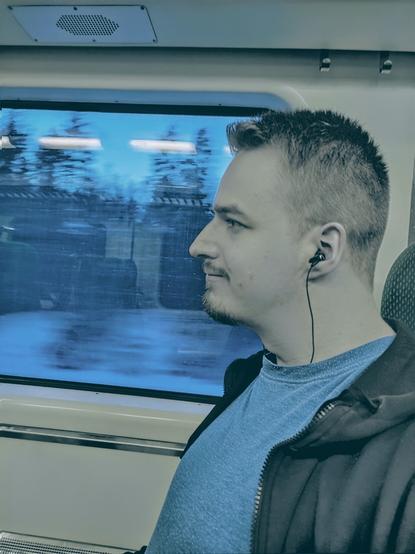 A man sitting on a chair in a train looking out the window with earphones on.
