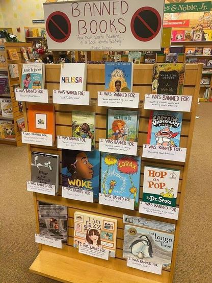 A wooden stand in a book store with the sign "Banned Books" then the quote, "Any book worth banning is a book worth reading - Isaac Asimov". They are all children's books and include, Melissa, "A Wrinkle in Time, Captain Underpants, The Lorax, I am Jazz, woke, The Lion, the Witch and The Wardrobe,  Hop on Pop.