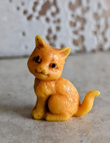 An image of a small plastic, orange cat in a sitting position, with its head turned and facing left, its tail pointing slightly upward.