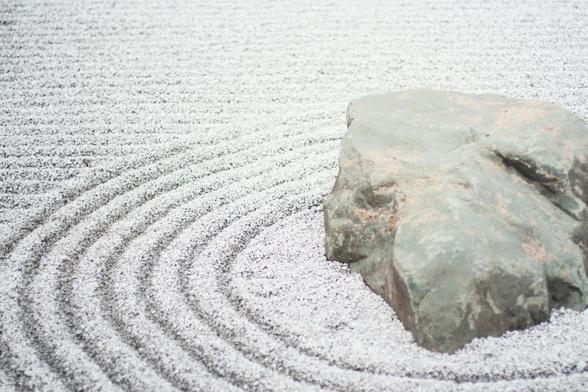 A stock photo by Kari Shea, via Unsplash.com, of a Japanese zen-style dry rock garden at The Richard & Helen DeVos Japanese Garden, Meijer Gardens, Grand Rapids, United States, featuring a large rock with raked, concentric circles in the sand.