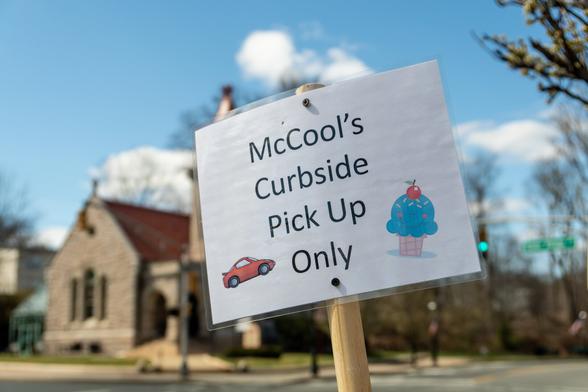 A color photograph of a sign on a wooden stick reading “McCool’s Curbside Pickup Only” featuring clip art of a red car and an ice cream cone with blue ice cream, a cherry on top, and a smiling face”.

In the out of focus background you can see blue skies, and a grey stone and reddish roofed Romanesque Revival building that once served as the town library, and now houses the Museum of Early Trades and Crafts.