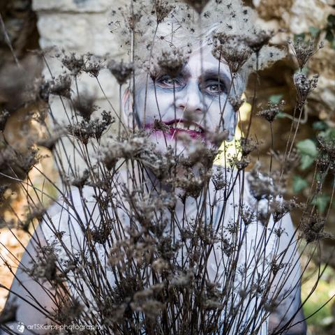 color photography of a woman in a white wedding dress holding a bouquet of dried flowers in front of her so that her smiling face is only partially visible.  Her head is adorned with fluffy, dry plant parts and her face is made up very pale, her mouth is painted dark red and extends beyond her actual lips on the left and right.  she is standing in front of an old stone arch surrounded by old woody tendrils and other vegetation.