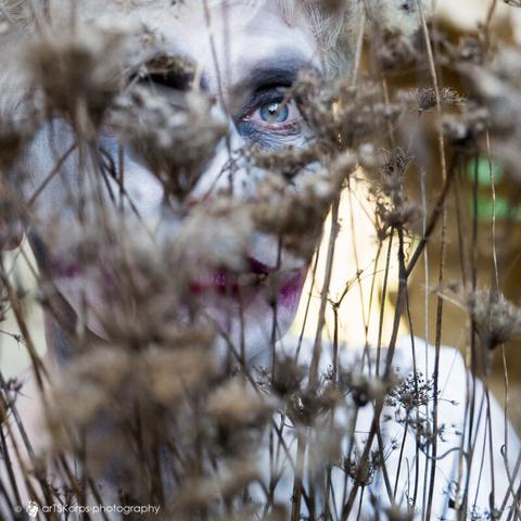 color photography of a woman (only the head can be seen in the photo) who is almost hidden behind many stalks with dried flowers.  Her head is adorned with fluffy dry plant parts and her face is made up very pale, her eyes are blue and the gaze that looks through the plants is intense.  Her mouth is painted dark red and extends beyond her actual lips on the left and right.