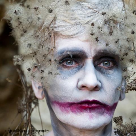 color photograph of a woman (only the head can be seen in the photo).  Her hair is white, her head is adorned with fluffy, dry parts of plants and her face is made up very pale, her eyes are painted blue and the gaze that looks through the plants is intense.  Her mouth is painted dark red and extends beyond her actual lips on the left and right.