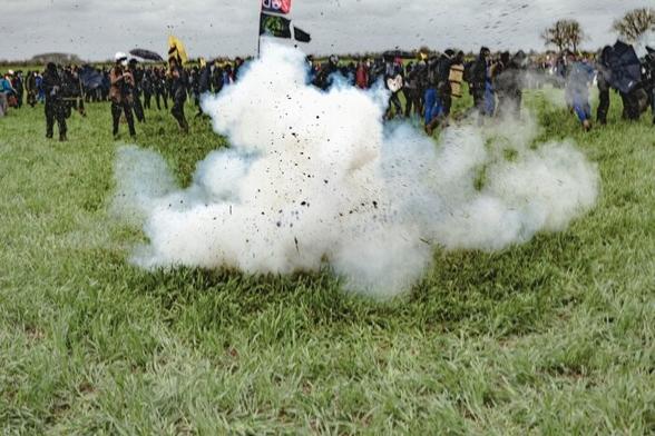 Image de la détonation d'une grenade en plein champs. Des morceaux de terre et de cailloux volent en toutes directions. 

Derriére on aperçoit des manifestants.