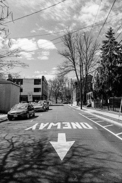 A black and white photograph looking down a side street in a business district from the middle of the road. Buildings line both sides, some cars are parked. A car is visible, driving from left to right on the cross street at the end of the road. Across from the end, a hill - actually the base for elevated train tracks - is visible.
Closest to the camera, the letters “ONE WAY” are painted in white on the road, with the arrow pointing towards the viewer at the center of the image.