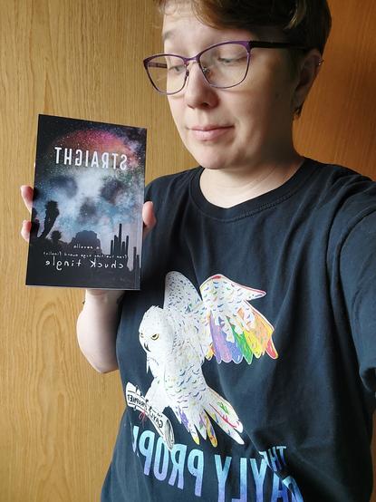 A white person with brown hair and purple glasses standing inside looking at a book in their hand. The book is called STRAIGHT by Chuck Tingle and has a skull made out of starlike lights in the sky. 
The person wears a black t-shirt with a big owl with rainbow feathers and a rolled up newspaper in its beak. Underneath it says "The Gayly Prophet"