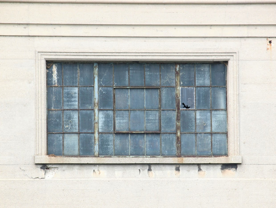 The building is beige and shows peeling paint and rust spots. Centred in the photo is a window with 11 panes across and 4 panes down, resembling a crossword puzzle with some light and some dark panes. There is no way to see through the panes due to dirt or something covering the windows. Rust is evident around all of the panes.