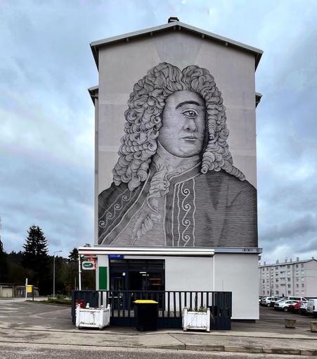 Streetartwall. On the outer wall of a four-story white house is sprayed / painted mural of a cyclops. In black and white and drawn in detail, a man with one eye and eyebrows in the middle of his face, looks over the city. He wears a powdered wig, a baroque vest and frills. A sympathetic cyclops.