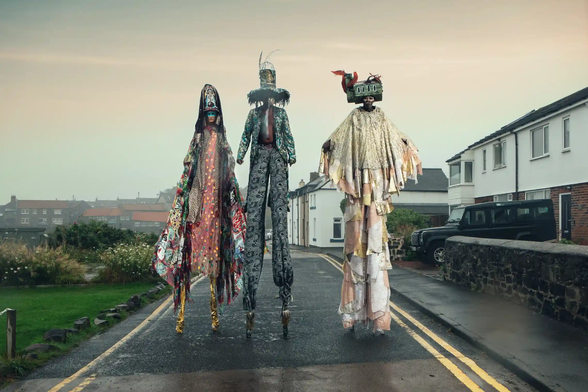 Photograph by Asiko, taken in Newcastle. It shows three people on very high stilts, in complicated, layered costumes made of still, colourful fabric, with bells and other ornaments attached. They have facepaints on and each is wearing a different hat, very elaborately made. In the background ordinary UK houses, street, plants, a landrover. It seems to have been taken early in the morning or at dusk. The whole atmosphere is surreal, otherworldly.