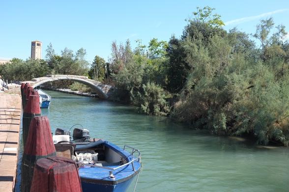 Ein Wasserweg auf der Insel Torcello (Lagune von Venedig). Motorboote am Kai zur Linken, rechts am Wasser Bäume und Büsche. Eine Steinbrücke. Hinten im Bild der Turm der Kirche Santa Fosca.
