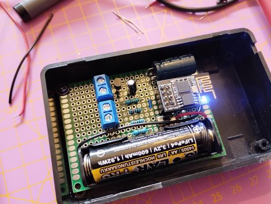 A soldered circuit board in a black plastic case. There's a LiFePO4 battery powering it. The LED of the ESP-01S is glowing. You can see a few resistors, capacitors, and a transistor.