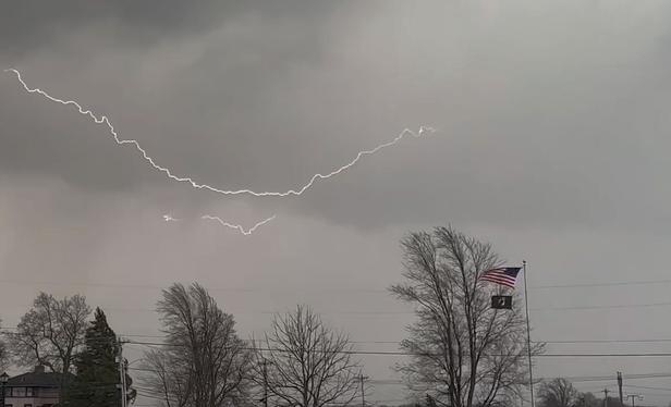 Thin, jagged lightning bolts, one spanning two-thirds of the image, are strung between the base of dark gray storm clouds. The shorter bolt of the two is partly obstructed by a rain curtain falling from the cloud. In the foreground a U.S. flag and a P.O.W. flag strain in the wind against the rope holding them to a flagpole.