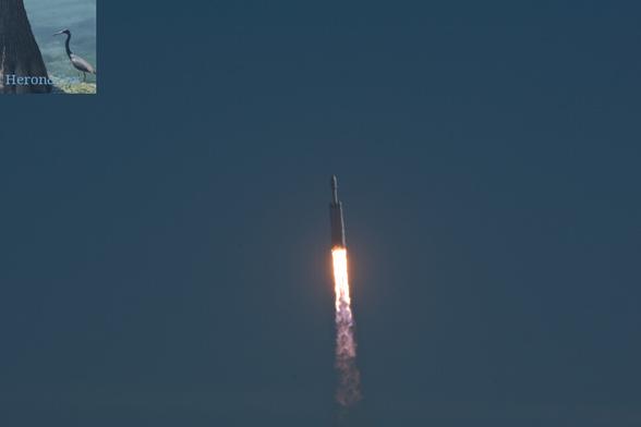 A photograph of SpaceX Falcon 9 Heavy rocket, with three white cylinders lined up side-by-side with the upper stage and fairing mounted atop the center core. Flames are emerging from the vehicle’s engines.
