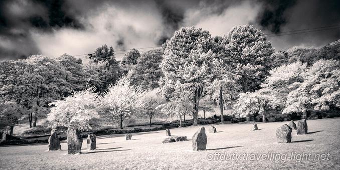 A small stone circle on sloping ground, with trees behind and clouds against a dark sky.