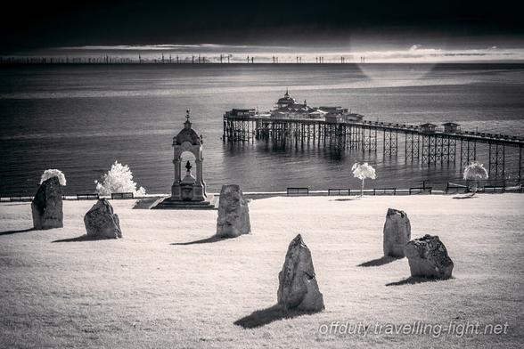 Part of a small stone circle in the foreground and a view out to sea with a seaside pier and an offshore windfarm on the horizon.