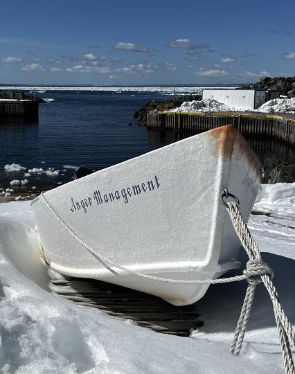 A snow-covered ocean wharf boat ramp with a white dory boat tied with thick white rope. The dory has a small amount of rust near the bow, with the  name "Anger Management" written in scripted letters. The sky is blue with a few puffy white clouds in the distance. The ocean bay is a deeper blue with small chucks of sea ice floating near the slipway, and masses of sea ice aggregated in the distance. Taken in  Bauline, Newfoundland and & Labrador (Canada) April 2023.