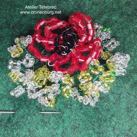 "Poppy" perlage object. A red flower is half surrounded by these small white-greenish flowers. The rocailles are very tiny, there is a needle for comparison.
