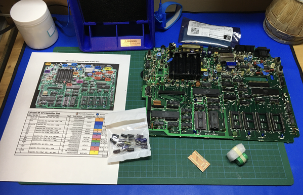 Electronics workspace with a mainboard of an Olivetti M10, documentation, a bag of replacement capacitors, a replacement memory battery, and a bare memory PCB.