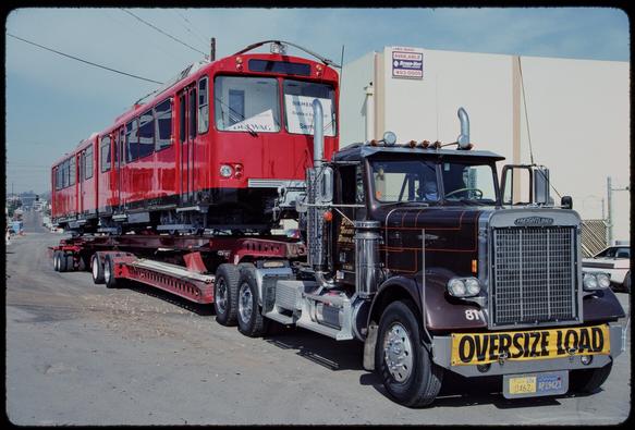 A red transit vehicle sits on a trailer behind a semi truck. A sign in the front right window of the vehicle reads "Siemens - Electrical equipment by Siemens." A sign in the front left window has fallen back and is largely unreadable, except the word "Duewag" at the bottom. A banner on the front bumper of the truck reads "Oversize Load."
