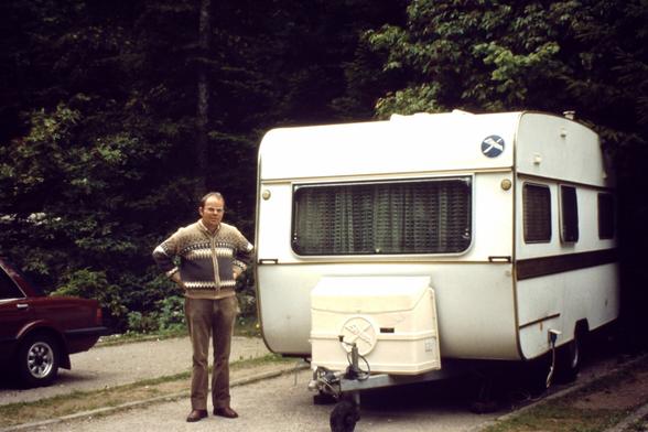 Ein Mann neben einem weißen Eriba Wohnwagen. Im Hintergrund eine "Wand" aus Bäumen.