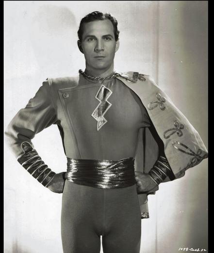 Medium close-up publicity still of Tyler dressed as Captain Marvel (Original Shazam character), has his fists on his hips , the short cape over his right shoulder. He looks directly at the camera with a calm resolute expression.