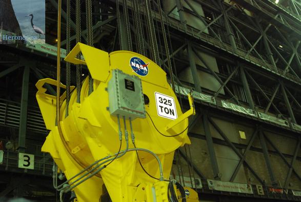 A photograph of a bright yellow pulley for a construction crane with the marking “325 TONS” and a NASA sticker against the background of truss-work that supports the VAB walls.