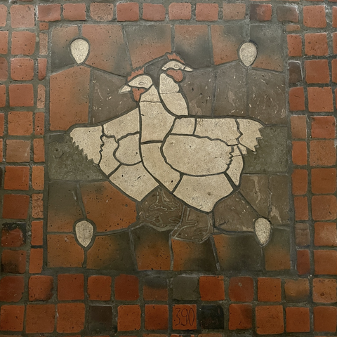 tile mosaic depicting two white chickens with red wattles and combs, on a square composition with an egg in each of the four corners, on red tile floor