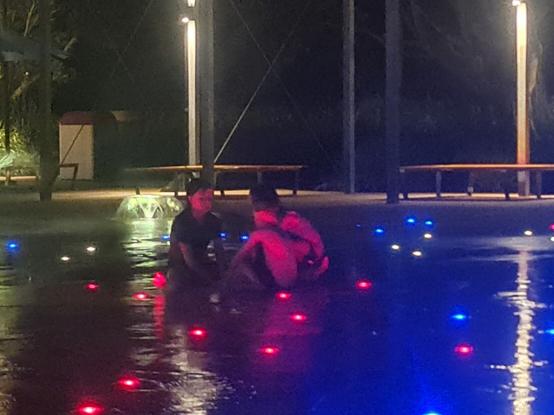 At night, 3 children sitting huddled together in the waterplay area, waiting for the fountains to restart.  They are lit by pink and blue ground lights.