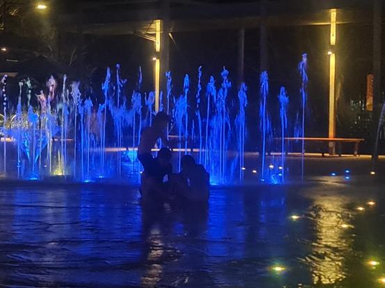 3 children playing at night in the waterplay area, they are silhouetted against blue lit fountains.