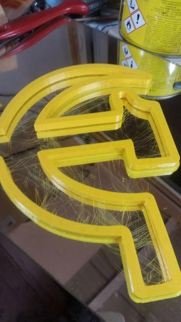 A hammer and sickle printed with yellow filament, with a lot of hairs along the rapid travels path.