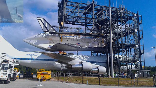 A photograph of Space Shuttle Orbiter Discovery. It is a plane-like glider with a white top and black heat-shield bottom. It is being lifted up inside a metal truss-work structure called the Mate-Demate Device. A Boeing 747 airplane has been moved inside the structure under the Shuttle, allowing Discovery to be mounted on the Shuttle Carrier Aircraft.