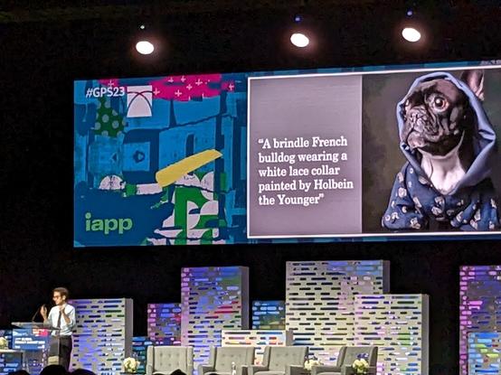 Alvaro Bedoya stands at a podium on the conference stage. A DALL-E illustration is displayed on a screen, with the prompt "A brindle French bulldog wearing a white lace collar painted by Holbein the Younger."