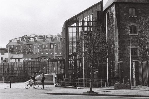 A grainy black and white photograph of the heineken brewery in Coek City, in landscape orientation. There are two small figures, a man and a woman in the front of the glass walled brewery entrance. City silouette can be seen on the background.