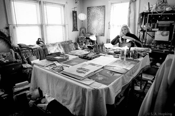 A black and white photo of a woman seated at her large desk. there are several sketch pads and lots of pens in front of her.