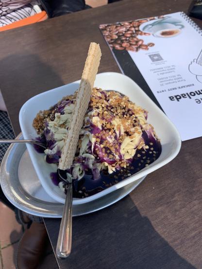 A picture of a bowl of tagliatelle-shaped pistaccio ice cream with blueberry sauce, crocant and a small waffle.
Next to it an ice parlour‘s menu, the picture done in half shade.