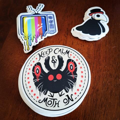 A photograph of three different vinyl sticker designs. Clockwise from top left: a melting CRT TV with a test pattern and a face with X-ed out eyes, a chibi style plague doctor that's just a head and a cape with no arms or legs, and a circular design of a cute Mothman surrounded by red stars and the words "Keep Calm & Moth On."