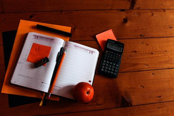 An open notebook, USB stick, calculator, pen, pencil, and apple, all on a dark wooden surface