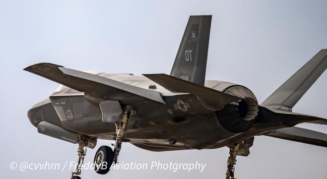 A closeup of a twin tailed all grey military aircraft is seen from the left lower corner and above. Its gear is down and "OT" can be seen stenciled on a tail. Just a hint of the cockpit can be seen but it is mostly obscured from this angle. The engines feathers are constricted showing a much more narrow opening as it drifts in to land