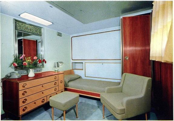 First class stateroom aboard the SS Michelangelo, 1965. There is a bed with very austere olive colored spread, a plain white wall, paneled closet doors, blocky modular furniture appropriate to the mid-1960s, and a chest of drawers, modernist style, with a vase of flowers on it. All the colors clash horribly.