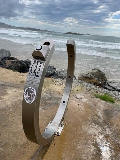 A silver, semi-circular horseshoe-type shape sits in front of a beach and coastline. It's a new style of dog wash with water jets to clean off pooches after their run on the beach.