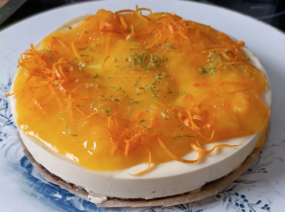 Die fertige Torte auf einer Tortenplatte.
Auf der weißen festen Creme ist eine orangene Sauce mit Orangenstücken verteilt. Darauf Limetten und Orangenzesten