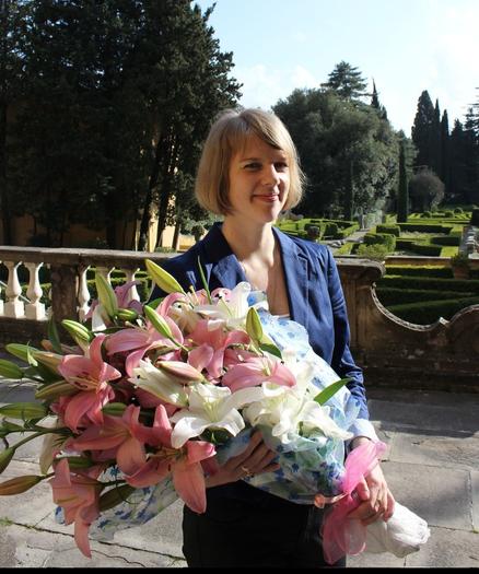 Newly minted PhD in blue jacket holding a large bouquet of lilys in front of garden