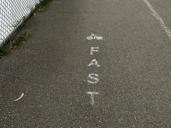 Photo of a bicycle symbol and the word word “FAST” stenciled on an asphalt-surfaced track. The photo was taken from a relatively close-up, oblique perspective. A small portion of chain link fence is visible to the left, and a portion of a painted lane separator stripe is to the right.