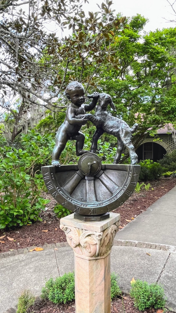 A statue of young boy holding up the leg of a lamb in playful way. Statue is on a pedestal, greenery on the background.