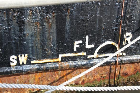 The side of a ship, near the water line. The upper part is painted black, with corrosion visible under the paint. The lower part is rusty. In the middle is the Plimsoll line mark, with "SW" and "F" markings for salt water and fresh.