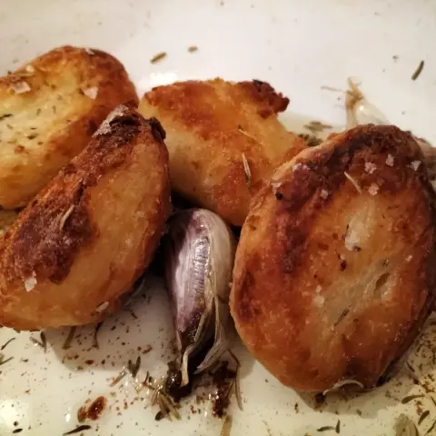 Four golden and crispy roast potatoes with garlic, rosemary and thyme in a white enamel roasting tin