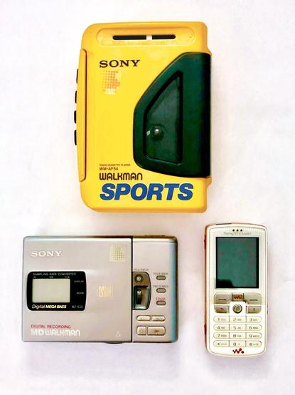 Three handheld electronic devices lying on a white backdrop. From the top, a yellow and black plastic cassette tape player from the 1980s; a silver metal MiniDisc player with an LCD screen from the 1990s; an orange and white plastic mobile phone with a colour TFT display from the 2000s.